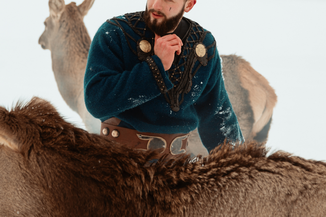 A Viking man wearing traditional clothing, standing with a deer in front of him and another deer behind him